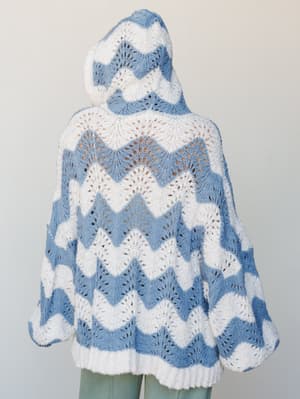 Shot from the back, this oversized hooded knit sweater has a dreamy, handmade quality — chevron wave stripes in cornflower blue and white work together in an open-stitch fan pattern that's equal parts cozy and collectible.
