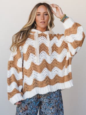 An oversized crochet-knit hoodie in a wavy chevron pattern — ivory and camel — worn with butterfly-print wide-leg pants and a stack of colorful beaded bracelets, giving off that perfectly lived-in, cozy-but-collected energy.