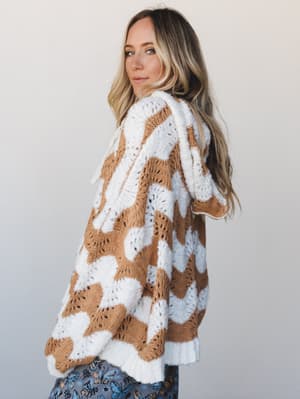 A model shot over the shoulder in a chunky wavy-knit hoodie in ivory and camel, the oversized silhouette and open-knit texture giving it that perfectly cozy, lived-in feel.