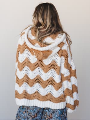 Shot from behind, a model wears a cozy oversized hoodie in a two-tone wavy knit pattern — ivory and camel alternating in scalloped chevron rows — layered over a blue butterfly print skirt, giving it that effortless mix-and-match boho energy.