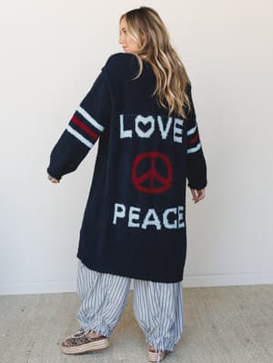Shot from behind, a model shows off a long, oversized navy boucle cardigan with 'LOVE PEACE' intarsia knit lettering and a red peace sign on the back, paired with wide-leg striped pants and espadrille platform sandals — cozy, expressive, and unmistak
