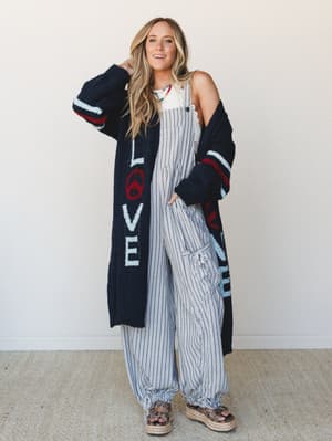 A long navy sweater cardigan with 'LOVE' lettering and a peace sign graphic is layered over wide-leg striped overalls and a white tank, styled with platform espadrille sandals and a colorful beaded necklace — the whole look has that relaxed, free-spi