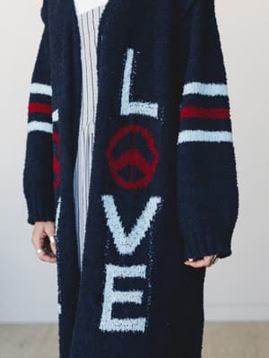 A cozy, oversized longline cardigan in navy with bold 'LOVE' lettering and a peace sign intarsia knit down the front, layered over a striped top — the kind of piece that says something without you having to.