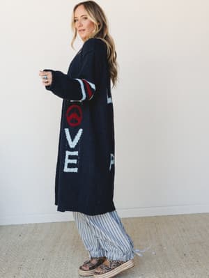 A long navy sweater cardigan with bold 'LOVE' lettering and a peace sign intarsia knit down the front panels, styled over pinstripe wide-leg pants and leopard platform espadrilles — the kind of outfit that has a personality before you even say a word