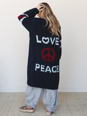 Shot from behind, a model with beachy waves wears a longline navy bouclé cardigan with 'LOVE' and 'PEACE' knitted across the back in powder blue, anchored by a bold red peace sign — paired with wide-leg striped pants and espadrille platform sandals f