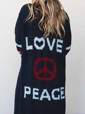 A cozy, oversized long cardigan in deep navy boucle knit, shot from the back — 'LOVE' and 'PEACE' spelled out in white intarsia lettering with a bold red peace sign in the center, worn by a model with wavy blonde hair against a soft neutral wall.