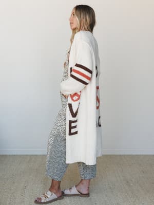 A long, cozy cream cardigan with bold letter graphic detailing and striped sleeves worn open over a floral wide-leg pant — the kind of layered, lived-in look you reach for without thinking twice.