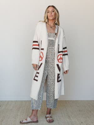 A long, oversized cream cardigan with 'LOVE' lettering and peace sign graphics layered over a grey leopard print jumpsuit — the kind of outfit that has a whole personality before you even leave the house.