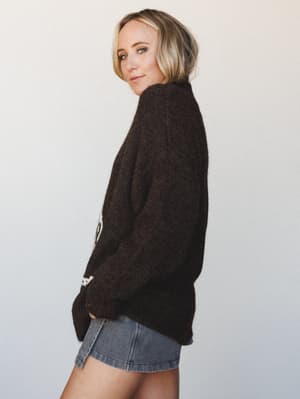 A side-profile shot of a model in a chunky dark chocolate fuzzy knit sweater with a subtle cream intarsia detail at the hem, paired with a denim mini skirt — the kind of sweater that makes you want to cancel plans and stay in, but in the best way.