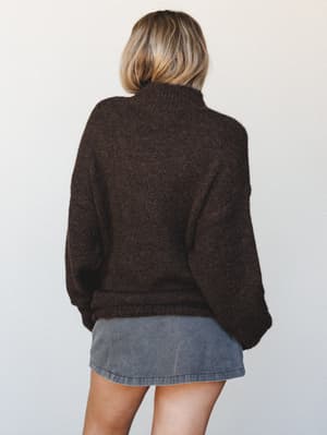 Shot from behind, a model layers an oversized chocolate brown knit sweater over a gray denim mini skirt — the kind of effortless fall combination that feels like it came together without trying.