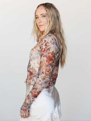 A floral mesh long-sleeve top in warm cream and dusty rose tones is styled with a white lace-trimmed bottom, shot in profile against a clean studio backdrop — soft, romantic, and effortlessly personal.