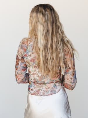 Shot from behind, a model with long wavy hair wears a sheer floral mesh long-sleeve top layered over what appears to be a satiny cream skirt — the back view quietly shows off the painterly floral print and the effortless, slightly undone feel of the