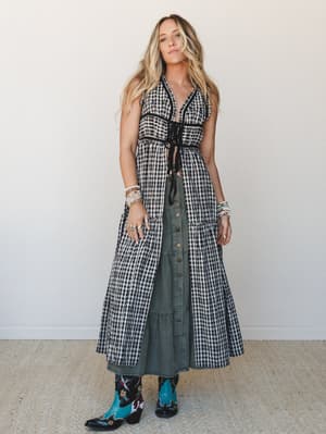 A layered boho outfit built around a black-and-white gingham maxi vest worn open over a sage green button-front midi skirt, grounded by embroidered teal cowboy boots — the whole look has that effortless music-festival-meets-farmers-market energy.