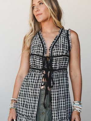 A black and white gingham vest with ruffle trim and a tie-front closure is layered over sage green wide-leg pants, styled with stacked beaded bracelets for a free-spirited, effortlessly put-together look.