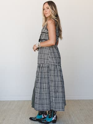 A black and white gingham maxi dress styled with statement turquoise embroidered cowboy boots and stacked beaded bracelets — shot in profile against a clean white wall with an easy, sun-soaked confidence.