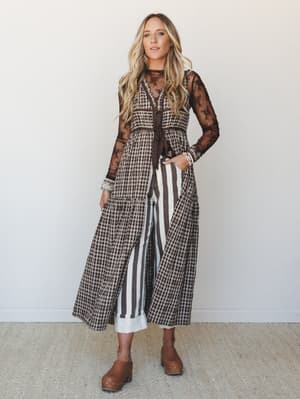 A layered, pattern-mixed outfit built around a chocolate gingham tiered maxi vest worn open over wide-leg striped pants, with a sheer lace long-sleeve top underneath — the kind of look that feels intentionally eclectic and effortlessly pulled togethe