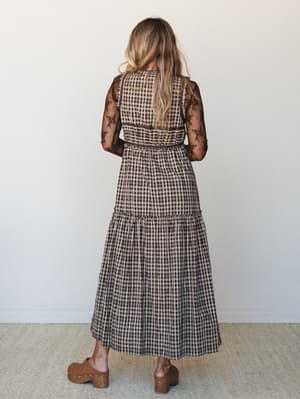 Shot from behind, a model wears a tiered gingham maxi dress layered over a sheer floral lace long-sleeve top, grounded with tan wooden clogs — the kind of effortlessly put-together look that feels vintage without trying too hard.
