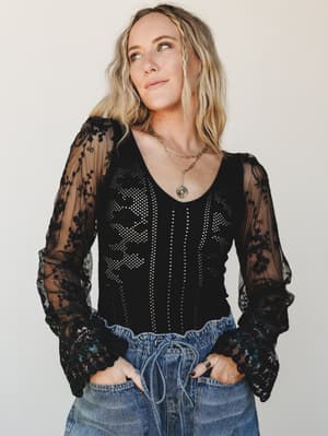 A black lace-sleeve bodysuit with sheer floral embroidered arms and a pointelle-knit body is styled with relaxed denim and layered gold necklaces — moody and romantic, but totally wearable.