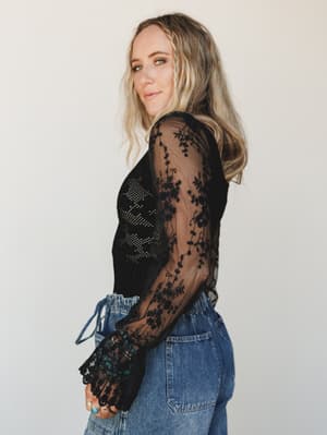 A floral embroidered mesh-sleeve bodysuit in black paired with relaxed denim, shot in three-quarter profile against a soft studio wall — the kind of outfit that reads equally well for a night out or a really good Saturday.
