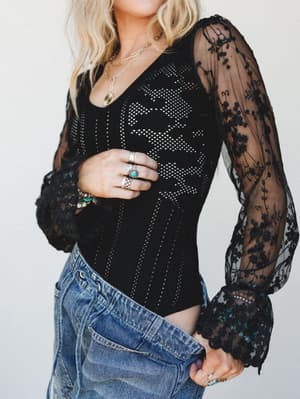 A black lace bodysuit with sheer floral-embroidered sleeves and a pointelle-knit body, styled with vintage-wash denim and layered jewelry for that effortless dark-romantic look.