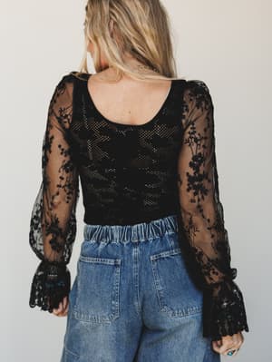 Shot from the back, this black lace bodysuit pairs sheer floral-embroidered sleeves with an open-knit bodice and flared cuffs — styled with relaxed wide-leg denim for an effortlessly moody, expressive look.