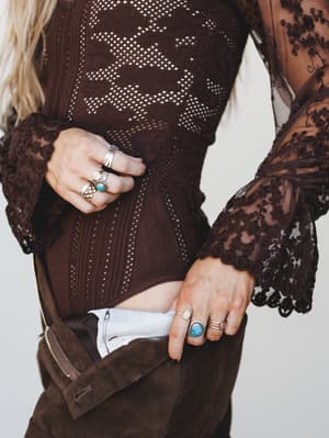 A close-up torso shot layering a dark brown pointelle-knit and lace bodysuit over wide-leg suede trousers, styled with stacked silver and turquoise rings that do most of the talking.