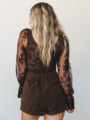 Shot from the back, this look pairs a dark brown floral lace bodysuit — sheer embroidered sleeves, open-knit body — with high-waisted olive-brown corduroy shorts, giving off a moody, vintage-boho energy that feels equal parts effortless and intention