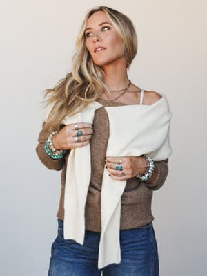 A relaxed, jewelry-forward look featuring a mocha knit sweater worn off one shoulder with a cream wrap draped across the body — stacked turquoise and stone bracelets, layered rings, and a delicate gold chain necklace do most of the storytelling.