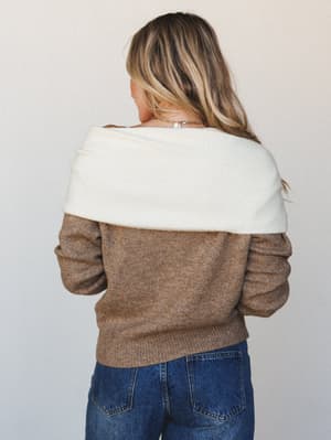 Shot from behind, a model in a mocha knit sweater with a dramatic oversized cream cowl folded off the shoulder, paired with dark indigo jeans — the kind of cozy layered look that makes you want to find a window seat.