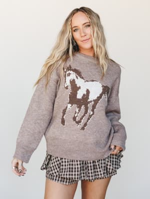 A relaxed, oversized knit sweater in warm sand with a bold intarsia horse graphic layered over a ruffled gingham mini skirt — the kind of outfit that feels effortlessly Western-leaning without trying too hard.