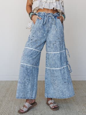Acid-wash wide-leg denim pants with eyelet embroidery and tiered paneling, styled with a white ruffled crop top, stacked beaded bracelets, and neutral slide sandals — casual boho with a lot of personality.