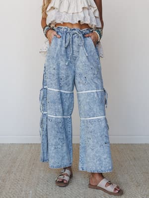 Acid-washed wide-leg denim pants with eyelet embroidery and tiered side ties, styled with a white ruffled crop top and sandals for an effortlessly boho warm-weather look.