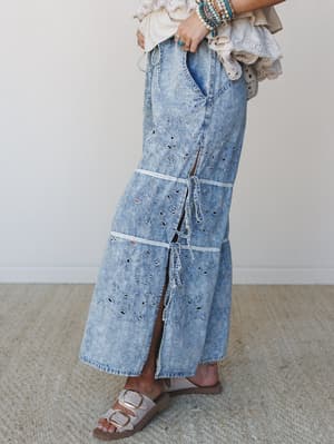 A side-angle shot of an acid-wash denim wide-leg pant covered in eyelet cutwork and tiered drawstring details, styled with a cream eyelet ruffle top, stacked turquoise bracelets, and neutral buckle sandals — boho-practical at its best.