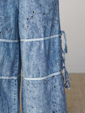 A close-up detail shot of acid-washed denim wide-leg pants covered in delicate floral eyelet embroidery, with ribbon-trimmed tier seams and a side tie closure — the kind of piece that makes you reconsider everything you thought about denim.