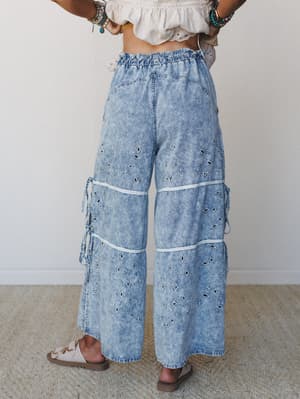 Shot from behind, these acid-washed wide-leg denim pants do a lot — eyelet embroidery, lace trim insets, side tie details, and a paperbag waist, styled with a cropped eyelet top and stacked bracelets for a full boho moment.