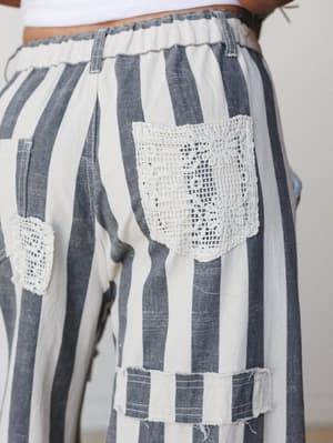 A close-up detail shot of wide-leg striped pants in charcoal and cream, featuring crochet lace patch pockets and raw-edge fabric appliqué — the kind of piece that looks like it has a whole story already.