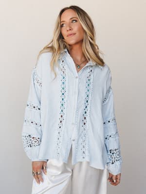 A breezy baby blue button-up top with intricate crochet lace insets along the front placket and sleeves, styled with white wide-leg pants and layered silver jewelry — effortlessly romantic and made for slow mornings that turn into something.