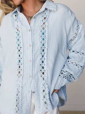 A close-up of a soft baby blue button-up top in gauzy cotton, with intricate floral crochet lace trim running down the placket and sleeves — the kind of top that makes a simple outfit feel intentional.