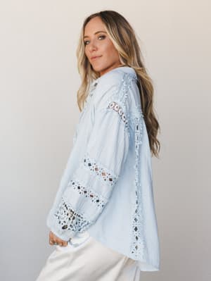 A soft, dreamy button-up top in baby blue gauze with floral crochet trim running down the sleeves and front — the kind of top that makes a Tuesday feel intentional.