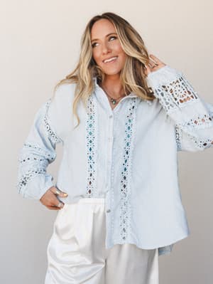 A light and airy button-up top in soft baby blue with intricate crochet lace trim down the front placket and sleeves, paired with white wide-leg pants — the kind of outfit that feels equally at home on a breezy patio or a weekend farmers market run.