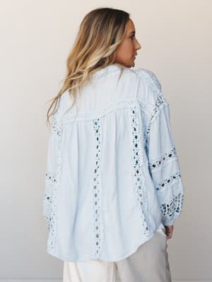 A flowy baby blue gauze blouse shot from the back, all soft volume and intricate lace inset panels — the kind of top that makes a casual afternoon feel a little more like something.