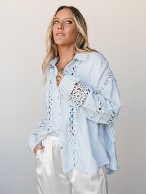 An oversized button-up top in soft baby blue with intricate crochet lace trim throughout — the kind of piece that looks like it belongs on a slow morning somewhere coastal, paired with white wide-leg trousers and layered turquoise jewelry.