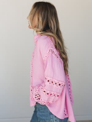 A side-profile shot of a long-haired model wearing a candy pink gauze button-up top with intricate crochet lace paneling and bell sleeves, paired with denim — soft, romantic, and very much the kind of top you buy and immediately want to wear everywhe