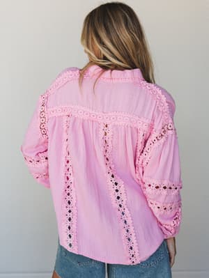 A back-view shot of a candy pink button-up blouse with intricate eyelet lace trim running vertically and horizontally across the body and sleeves, paired with light wash denim shorts — soft, feminine, and full of handcrafted detail.