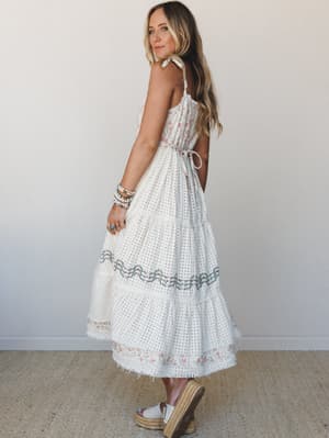 A cream tiered maxi dress with delicate floral embroidery and polka dot texture, styled with espadrille platform sandals and a stack of boho bracelets — shot from behind with a glance-back pose that shows off the open back and tie closure.