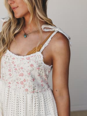 A close-up of a cream floral sundress with tie shoulder straps and eyelet skirt, styled with a mustard bralette peeking through and a turquoise pendant necklace — the kind of outfit that makes a farmers market feel like a whole moment.