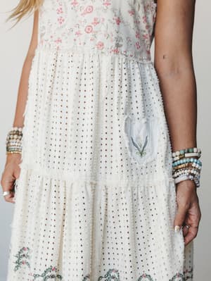 A close-up detail shot of a cream maxi dress with mixed floral print bodice and eyelet tiered skirt, styled with stacks of beaded bracelets on both wrists — the kind of dress that feels like it has a whole story already.
