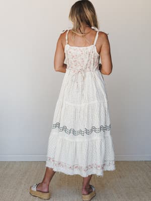 A cream midi-maxi dress shot from the back, showing off its tiered eyelet skirt, floral embroidered bodice, and delicate shoulder tie straps — the kind of dress that makes a farmers market feel like a special occasion.