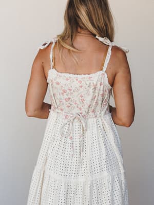 A back-view shot of a flowy cream maxi dress with a floral-print bodice, eyelet tiered skirt, and tie shoulder straps — the kind of dress that feels equally at home at a garden party or a slow morning at the farmers market.