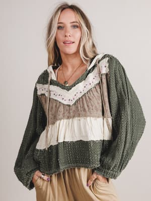 Woman wearing a layered outfit with a green cardigan over a patterned top and beige pants on a plain background
